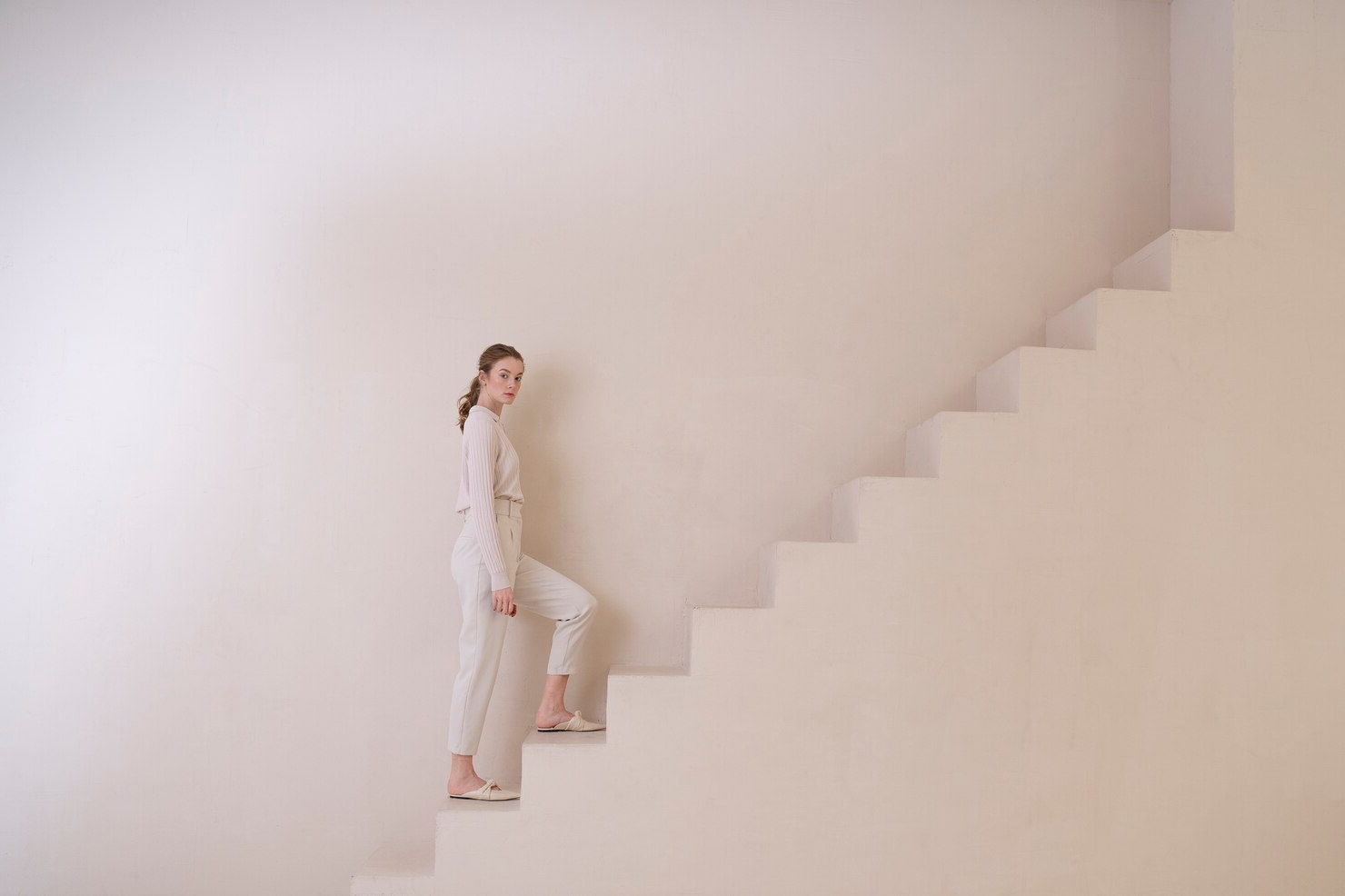 Person in light-colored clothing standing on a minimalist staircase, appearing to ascend. The environment is clean and modern with soft, neutral tones, creating a calm and serene atmosphere.