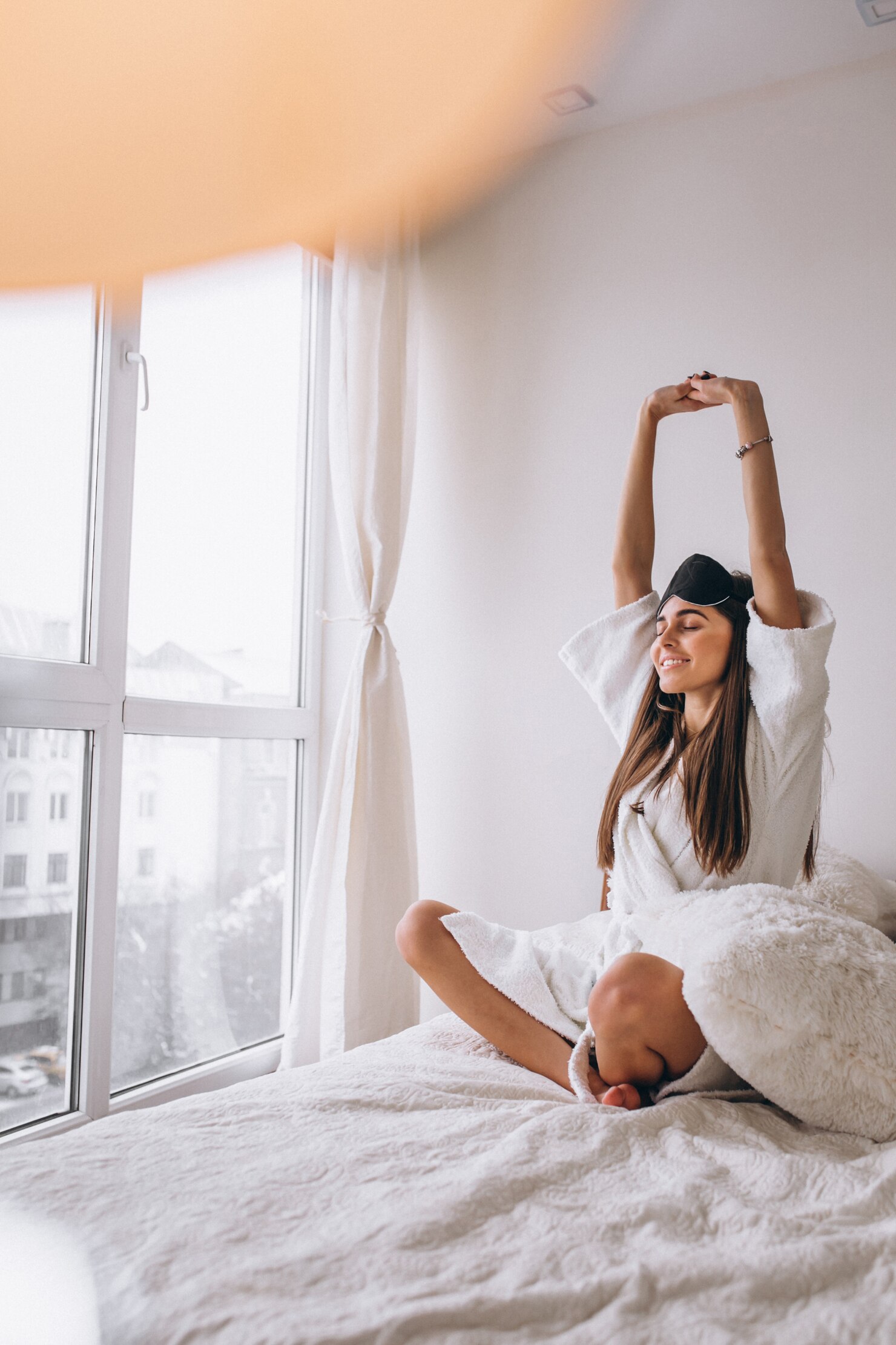 Person in a white robe sitting cross-legged on a bed, stretching arms overhead in a relaxed morning pose. The room is bright with natural light streaming through large windows, featuring white curtains and soft bedding, creating a calm and peaceful atmosphere.