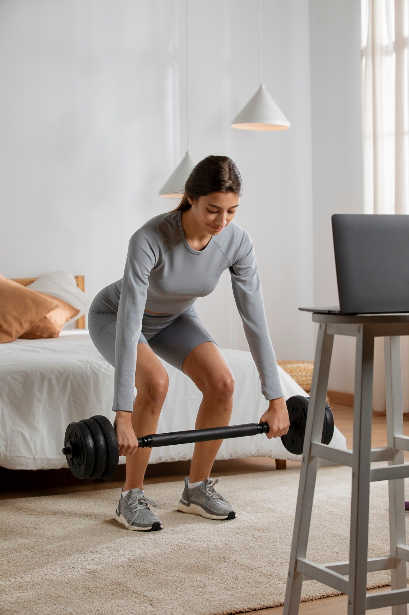 Woman in light gray athletic wear performing a deadlift with a barbell in a cozy bedroom. She is positioned near a bed with white bedding and a laptop on a tall table, suggesting she may be following an online workout. The room is softly lit with natural light and decorated in neutral tones, creating a calm and focused atmosphere.