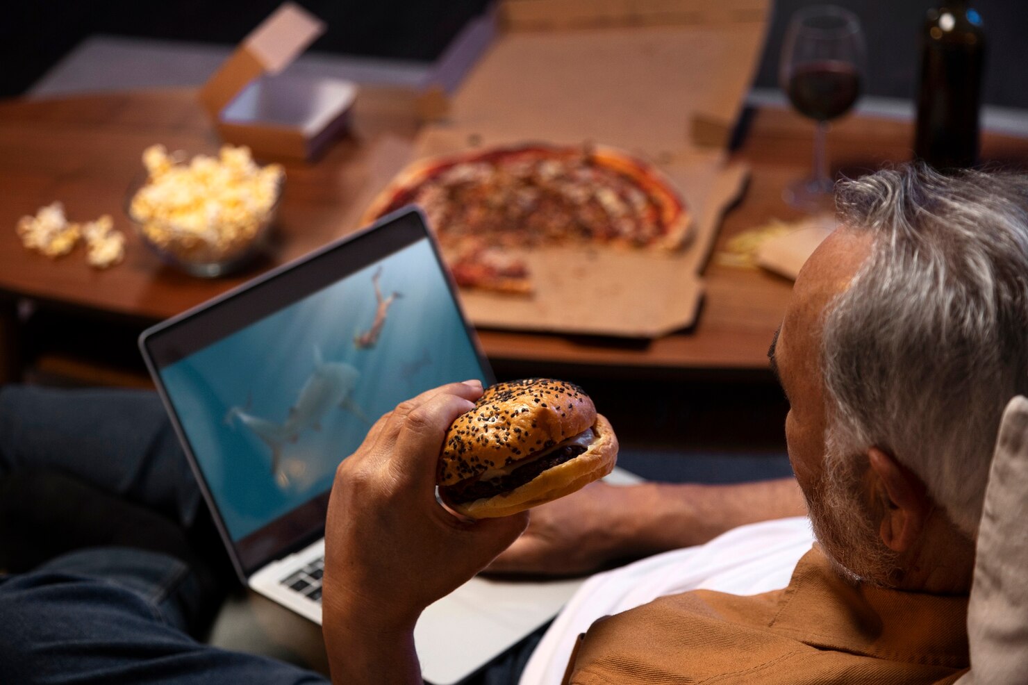 Man with gray hair sitting on a couch, holding a hamburger in one hand and watching a laptop screen displaying an underwater scene with sharks and swimmers. On the table in front of him are a pizza in an open box, a bowl of popcorn, a glass of red wine, and a bottle. The setting is cozy and relaxed, suggesting a casual evening at home enjoying food and entertainment.