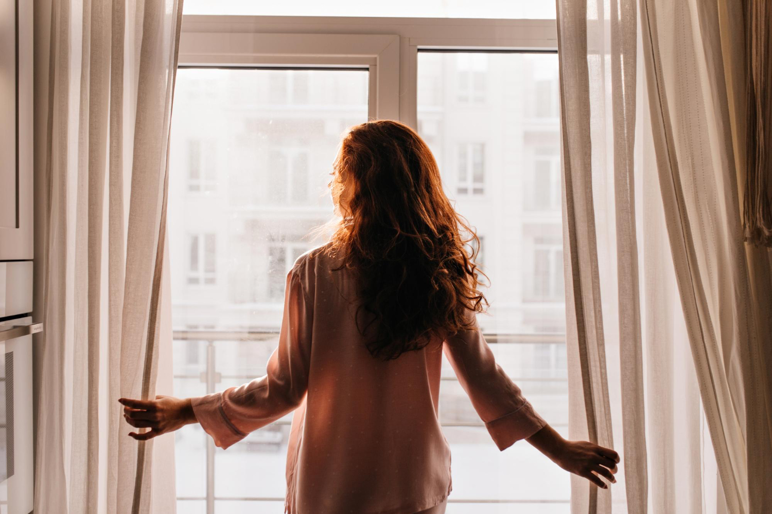 A woman stands at a large window pulling back sheer curtains as warm morning sunlight fills the room from behind her.