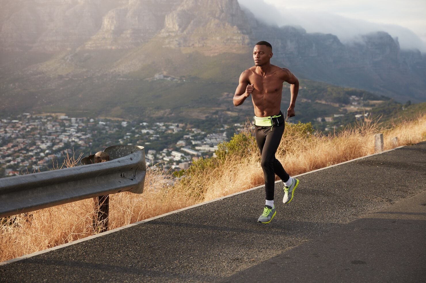 A shirtless man runs uphill on a paved road above a city, with dry grass and large mountains rising in the background.