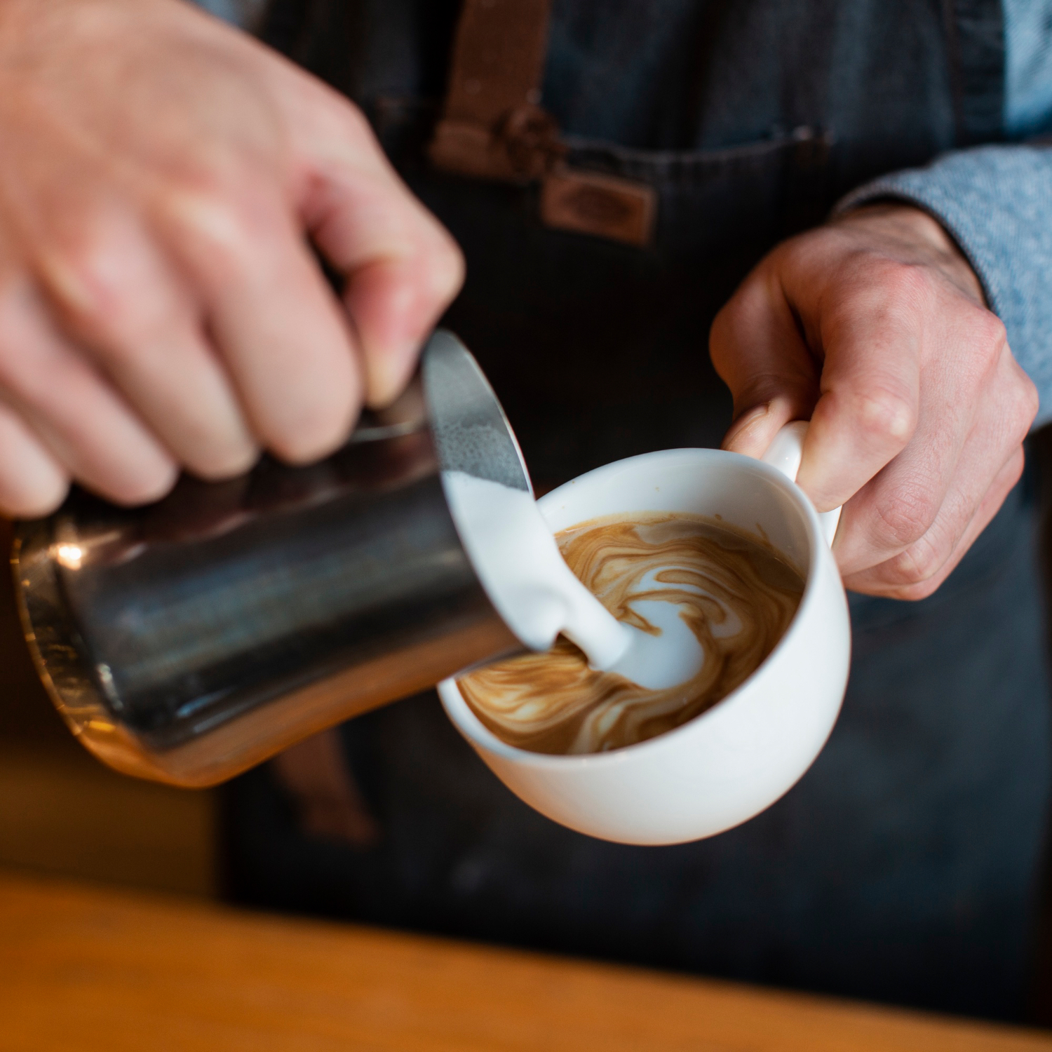 A close-up shows milk being poured from a metal pitcher into a white cup of coffee, creating swirling latte patterns.