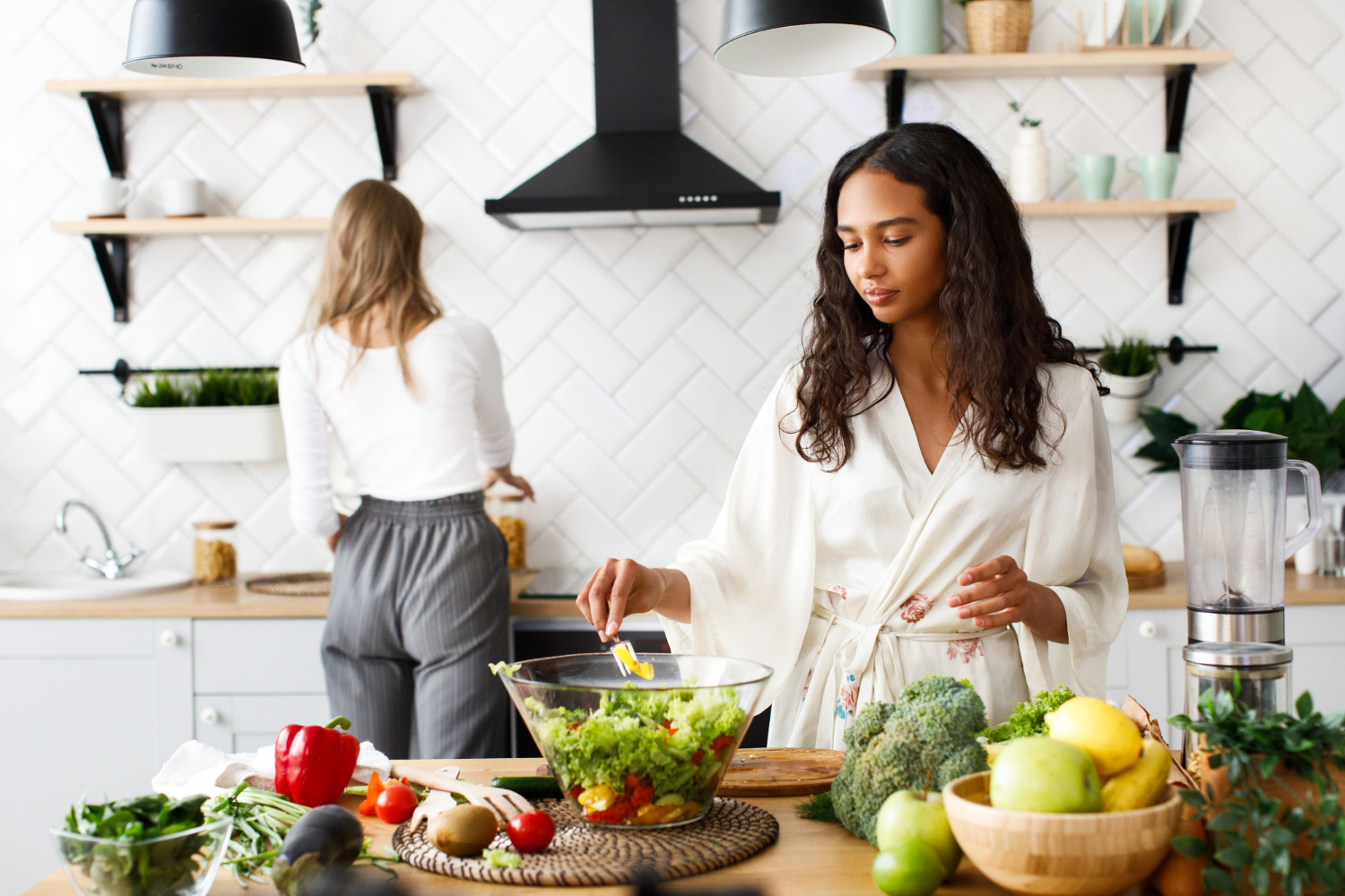 A woman in a light robe prepares a large salad in a bright kitchen while another woman works at the counter in the background. Fresh vegetables and fruit are spread across the wooden island.