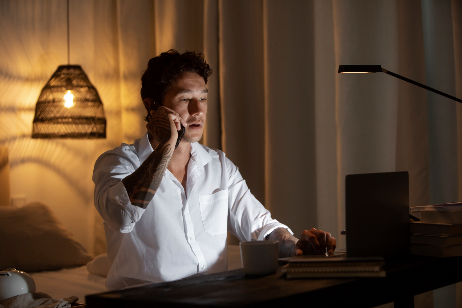 A man sits at a desk in a dim bedroom, talking on his phone while looking at a laptop screen. A mug, stacked books, and warm lamp light surround him late at night.