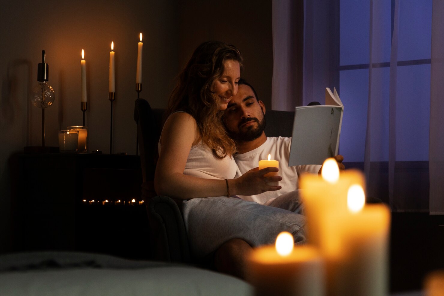 A couple sits together in a dark room lit by candles, reading a book in an armchair near a window with blue evening light outside.
