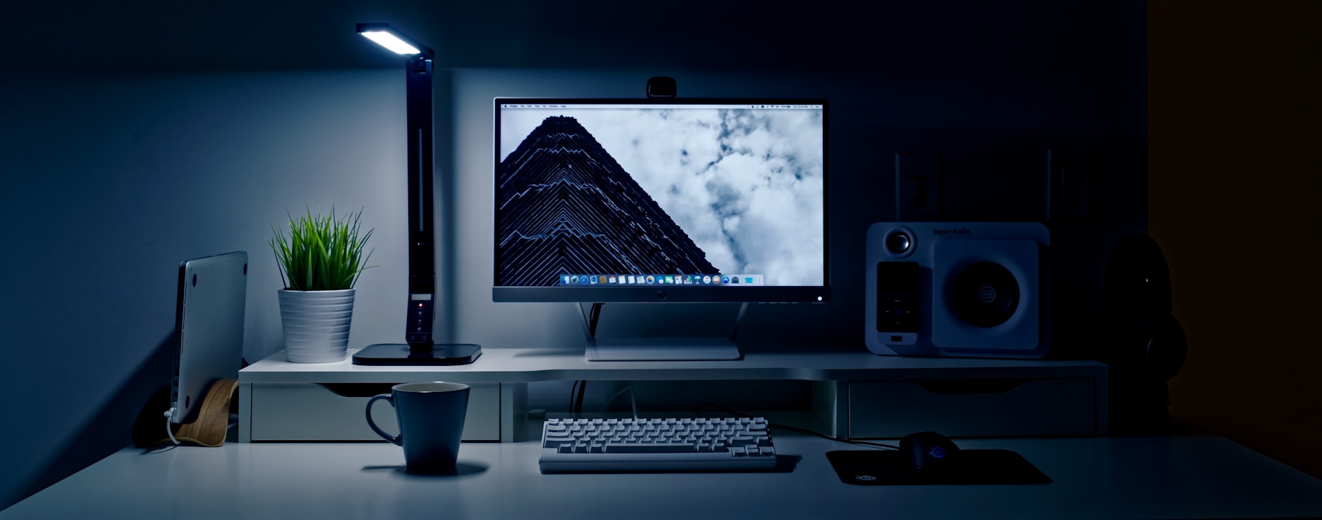 A desktop setup in a dark room with a glowing LED desk lamp, a computer monitor displaying a cloudy sky, a potted plant, a coffee mug, and tech accessories illuminated by cool blue light.