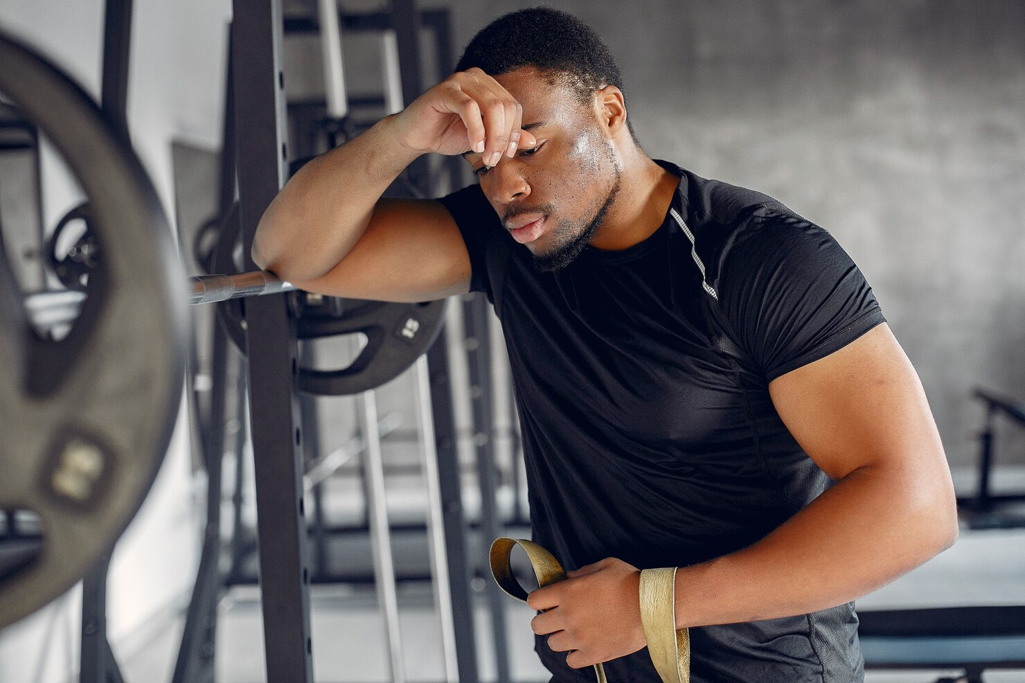 A sweaty man leans against gym equipment after a hard workout, looking overheated and fatigued.