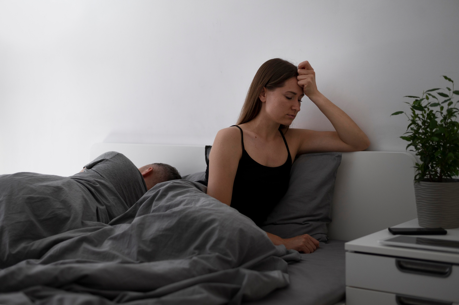 A sleepless woman sits on the edge of her bed at night beside a glass of water and a food journal on the nightstand.
