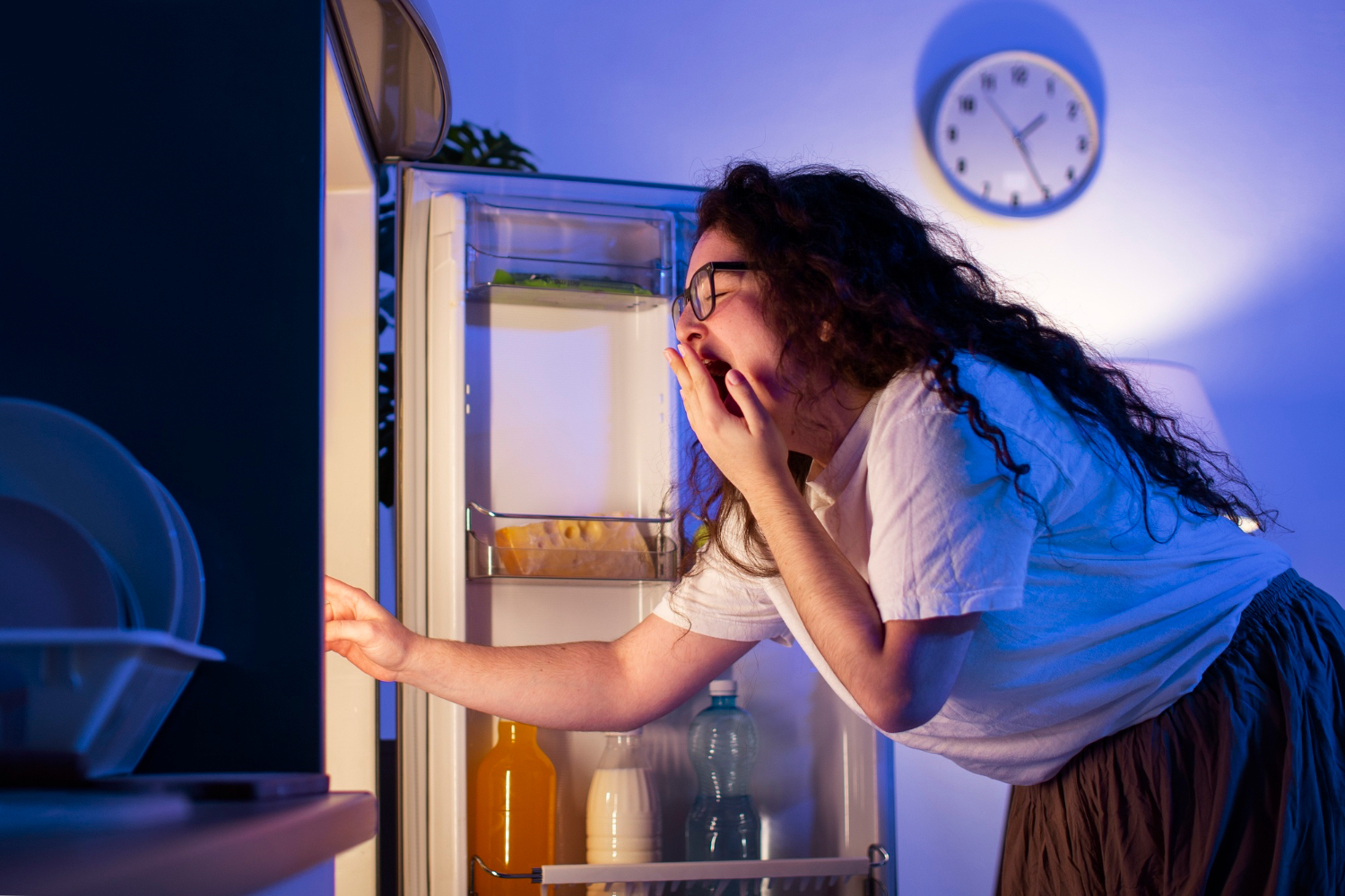 A sleepy woman stands at an open refrigerator late at night, lit by the fridge glow beneath a wall clock, suggesting bedtime hunger and a disrupted wind-down.