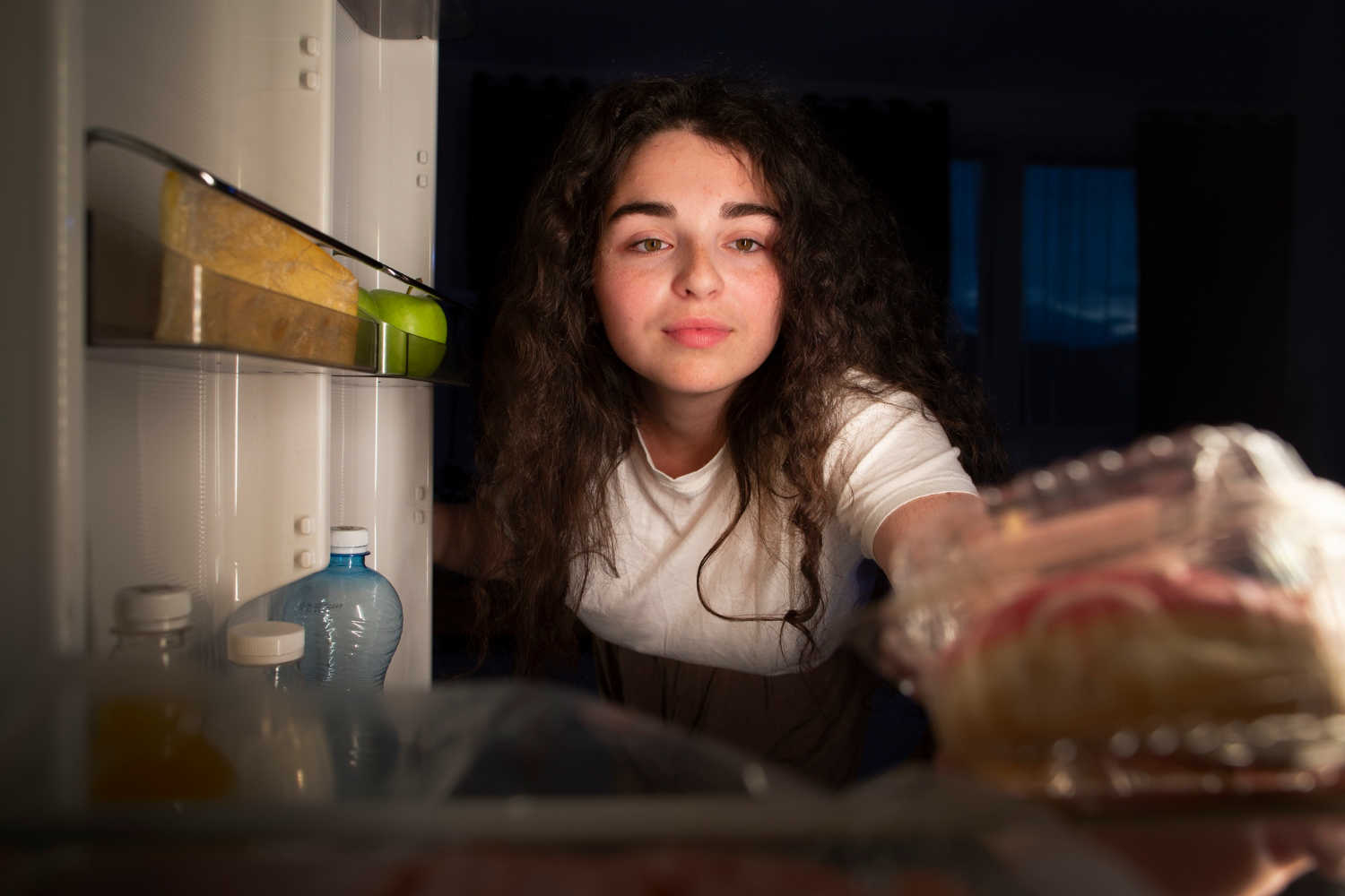 A woman reaches into an open refrigerator at night, illustrating the restless search for food that can happen when bedtime hunger becomes hard to ignore.
