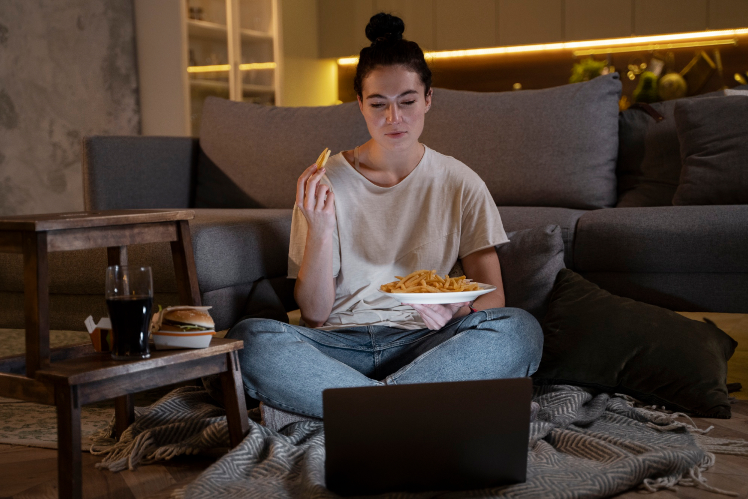 A woman sits on the floor late at night with fries, a sandwich, and a laptop, illustrating how casual evening eating can turn into a regular bedtime habit.