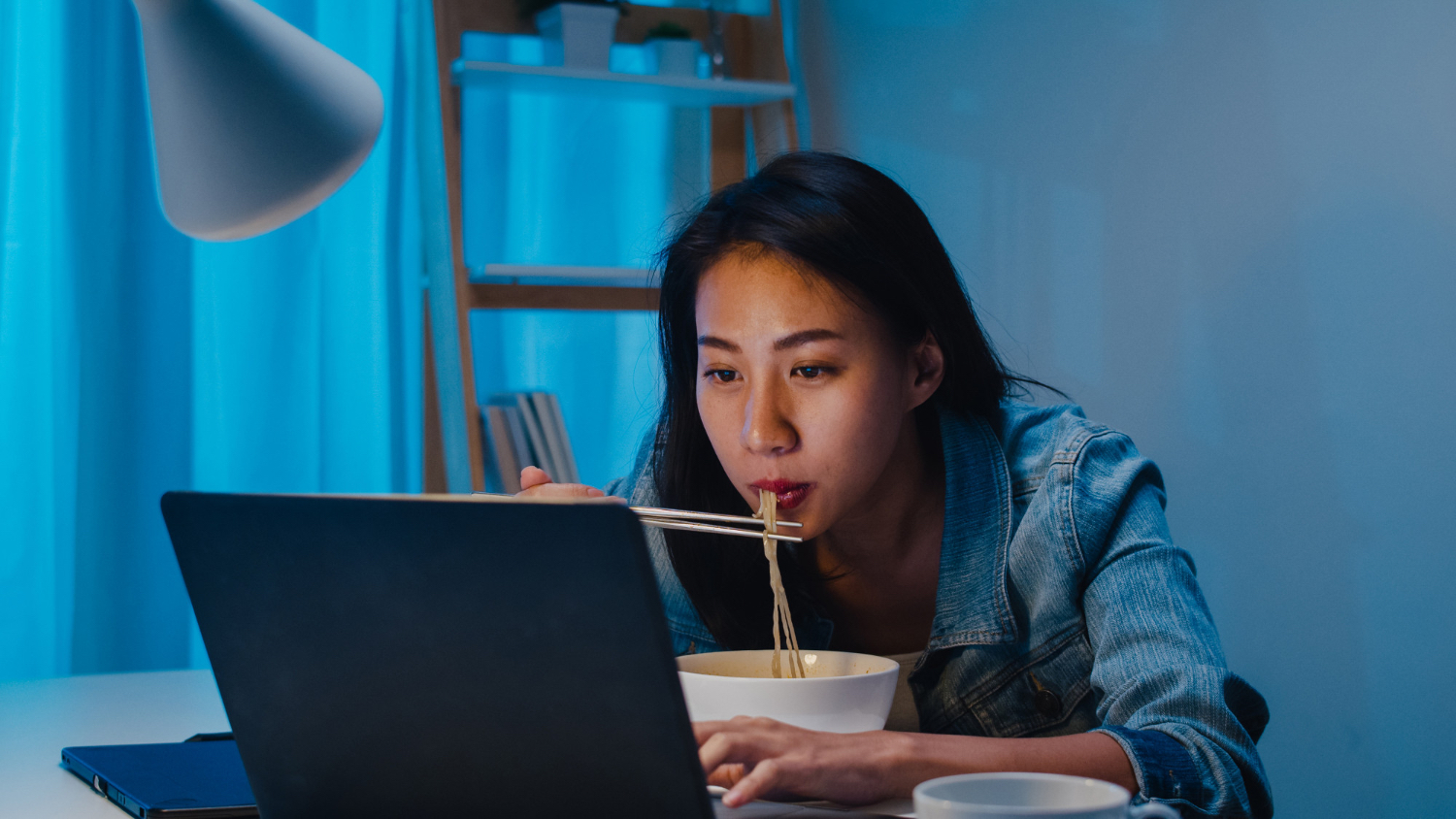 A woman eats noodles while leaning over a laptop in cool blue evening light, showing how late screen use and food can blend into a stimulating bedtime pattern.