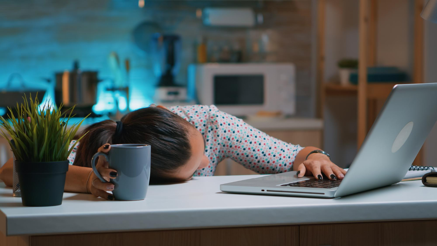 A person wearing a patterned shirt sits at a kitchen counter with their head resting on the surface, one hand holding a coffee mug and the other on a laptop keyboard. The kitchen is softly lit with blue accent lighting, and a potted plant sits nearby. The scene conveys a sense of exhaustion or overwhelm, suggesting late-night work or fatigue in a home environment.