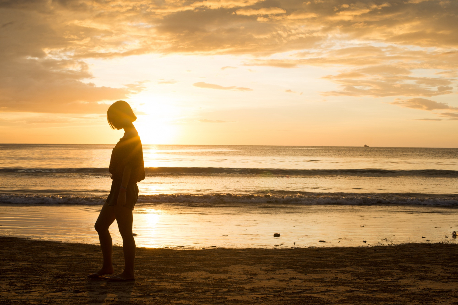 Silhouette of a woman with short hair walking along a sandy beach at sunset, head slightly bowed and arms relaxed at her sides. Gentle waves roll onto the shore and the sky is filled with warm golden and orange hues, creating a peaceful and contemplative atmosphere.