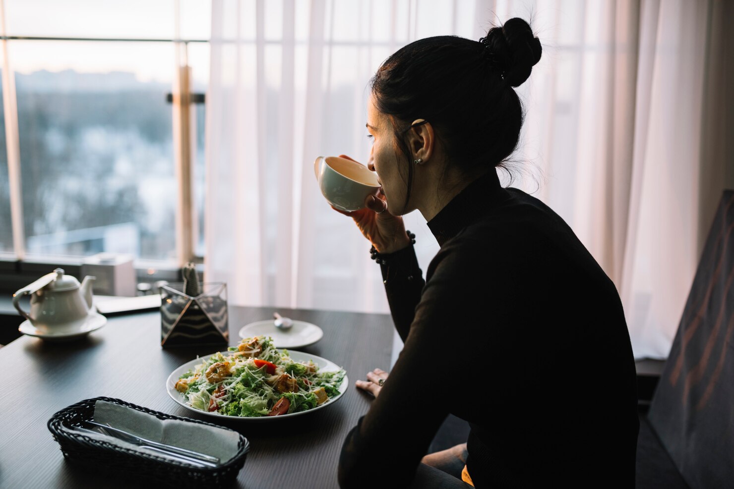 A woman with dark hair in a bun sits at a table by a window, drinking from a cup. In front of her is a large plate of salad with greens, tomatoes, and croutons. The table also holds a teapot, a napkin basket, and a small dish. Soft daylight filters through sheer curtains, creating a calm and peaceful atmosphere, suggesting a quiet meal or moment of relaxation.