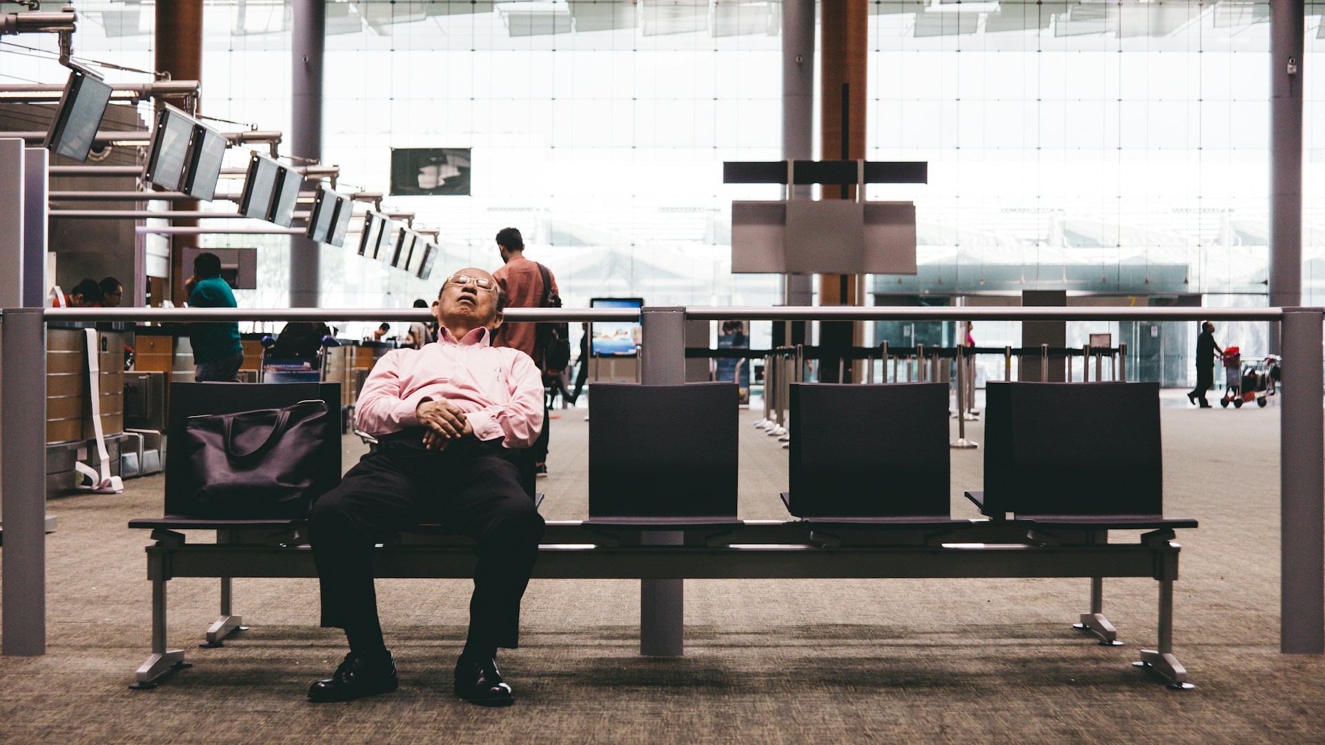 A person in business attire sits alone on a row of black chairs in an airport terminal, hands folded and legs stretched out, with a large black bag beside them. The spacious terminal features high ceilings, large windows letting in natural light, and several people in the background moving through the area. The atmosphere feels quiet and contemplative, suggesting waiting or anticipation.