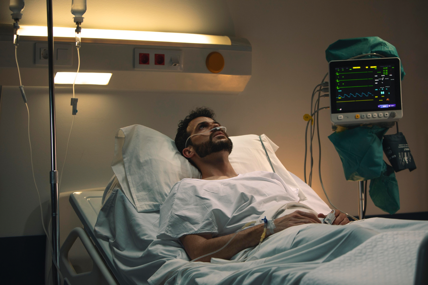 A person lies in a hospital bed wearing a white gown, with an IV attached to their arm and medical monitoring equipment nearby displaying vital signs. The room is dimly lit with clinical lighting, and the overall atmosphere feels serious and somber, suggesting a medical situation or recovery.