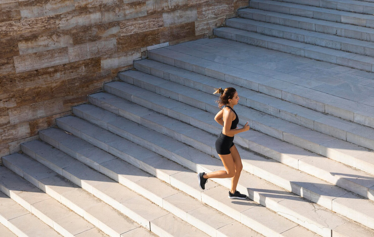 Woman in athletic wear running up a wide outdoor stone staircase, mid-stride, with sunlight casting shadows across the steps. The surrounding environment is clean and modern, with a textured stone wall to the side, creating an energetic and focused atmosphere.