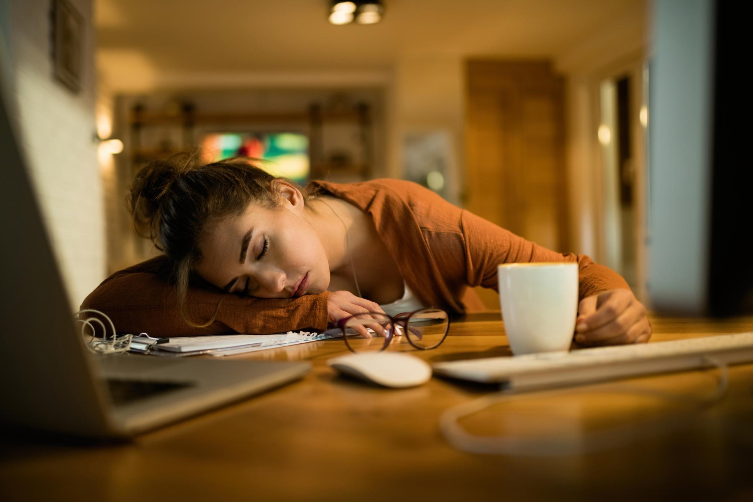 A woman has fallen asleep at a wooden table beside a laptop, coffee mug, glasses, and office items, suggesting exhaustion building into the evening.