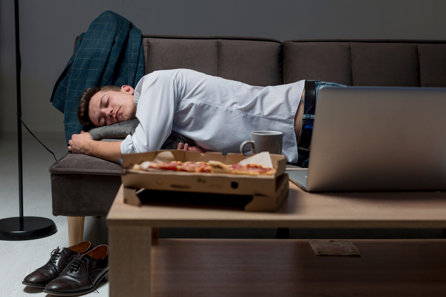 A man in work clothes has fallen asleep on a couch after dinner beside an open laptop, a coffee mug, and a pizza box, suggesting an accidental early-evening crash before bedtime.