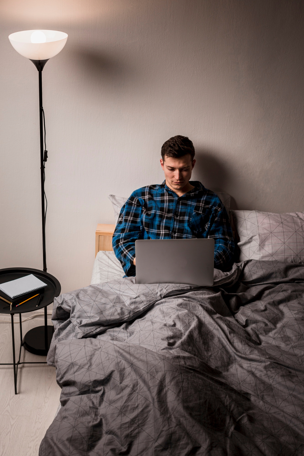 A man sits up in bed at night using a laptop under a lit floor lamp, illustrating how screens and room light can extend wakefulness after an evening nap.