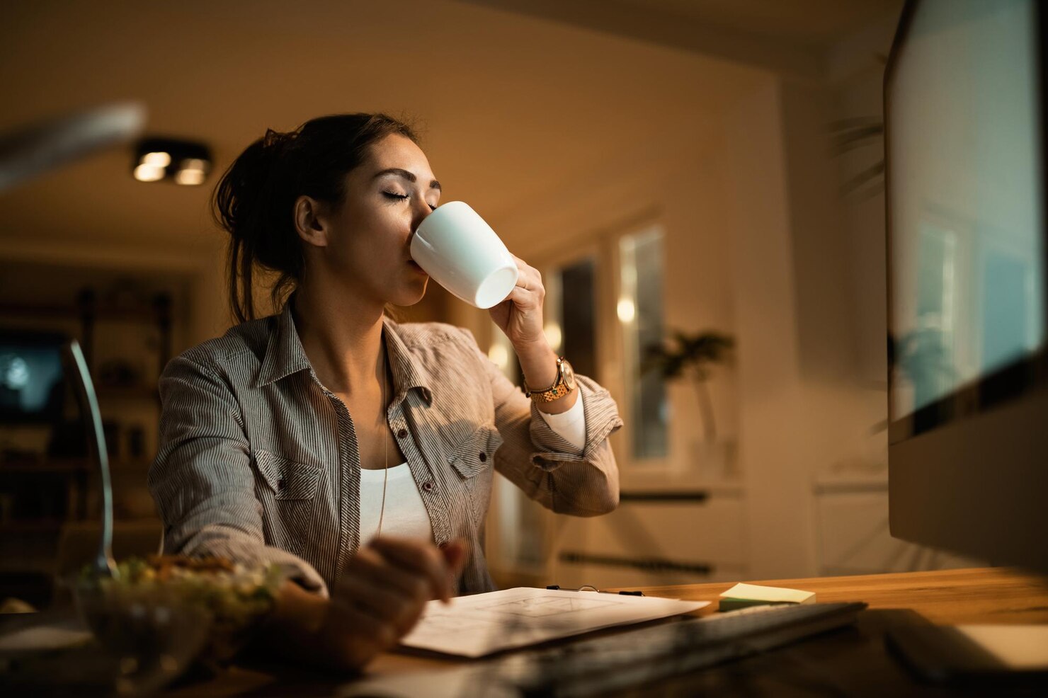 A woman sits at a desk at night, drinking from a white mug while facing a large computer screen. A clipboard and bowl sit on the desk in front of her in the warm indoor light.