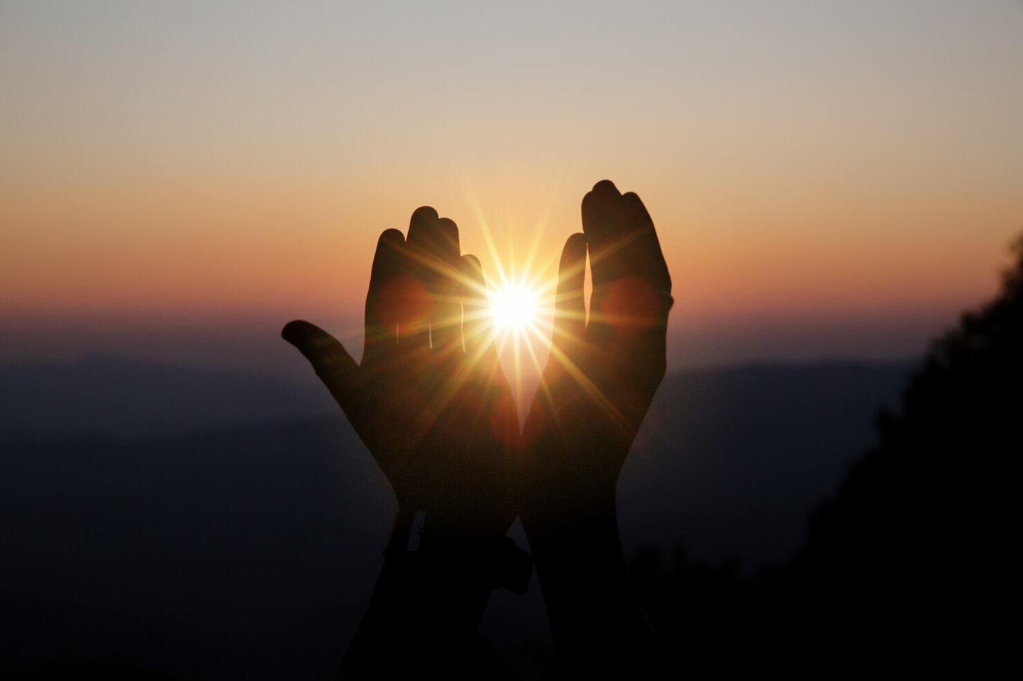Silhouette of two hands held up against a sunrise or sunset, with fingers slightly spread and the sun shining brightly between them. The background features a gradient sky transitioning from deep orange to soft blue, with distant mountains or hills visible in the lower part of the image. The scene conveys a sense of hope, warmth, and tranquility.