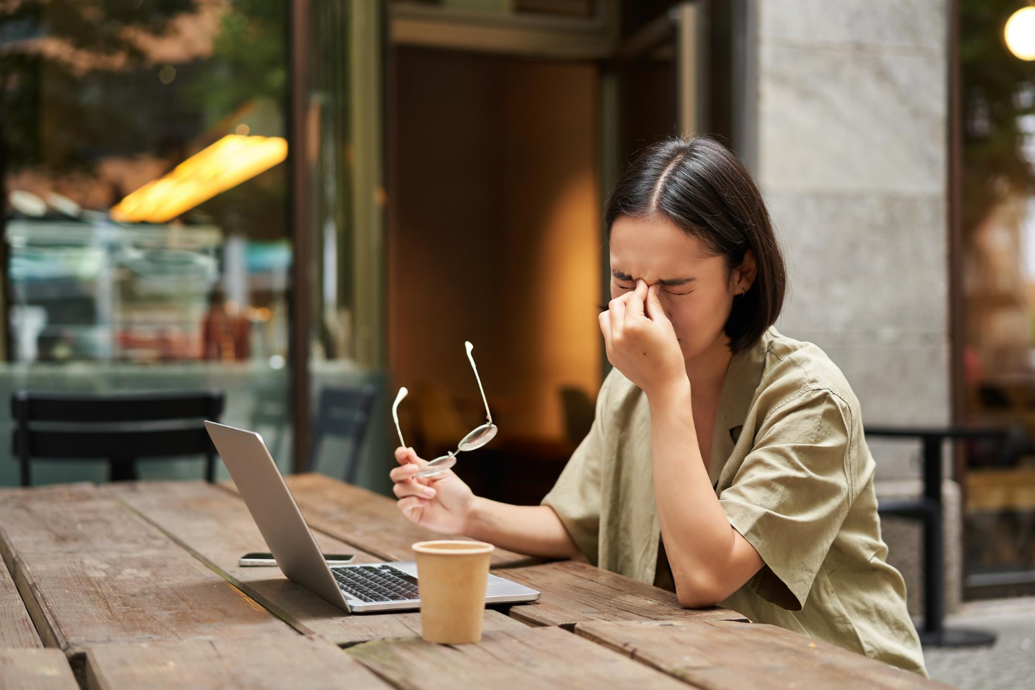 Person with short dark hair sitting at a wooden outdoor table in front of a laptop, holding eyeglasses in one hand and resting their head on the other. A disposable coffee cup sits on the table. The setting is a cafe with large windows and a warm, relaxed atmosphere, suggesting a moment of contemplation or focus while working or studying.