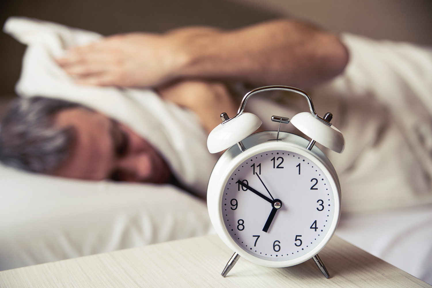 Analog alarm clock showing 6:50 on a bedside table in the foreground, with a person lying in bed in the background covering their head with a pillow. The room is softly lit, creating a sense of early morning reluctance or tiredness.