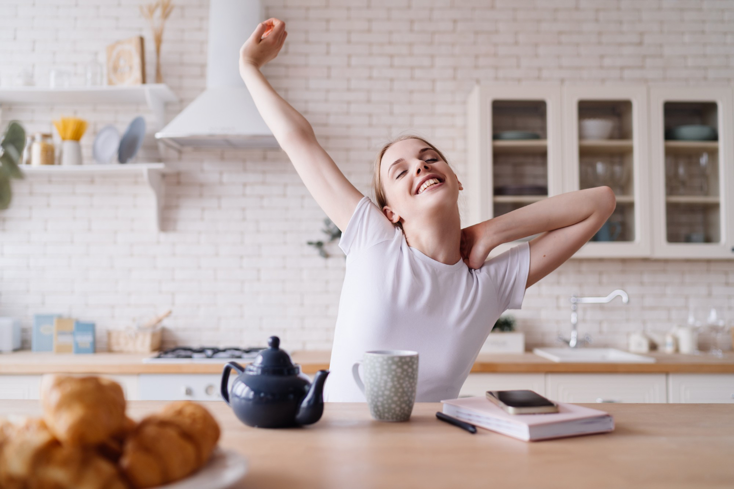 Person in a white t-shirt sitting at a kitchen table, stretching with one arm raised and the other bent behind their head. On the table are a teapot, a mug, a plate of croissants, a notebook, a pen, and a smartphone. The kitchen is bright and modern with white cabinets, open shelves, and natural light, creating a calm and relaxed morning atmosphere.