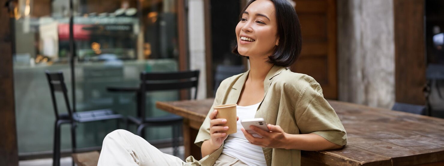 Person with straight dark hair sitting at a wooden outdoor table, holding a disposable coffee cup in one hand and a smartphone in the other. The person is dressed in light-colored casual clothing and appears relaxed, possibly taking a break or enjoying a quiet moment. The background features empty chairs and large windows, suggesting a calm cafe or restaurant setting with a peaceful and laid-back atmosphere.