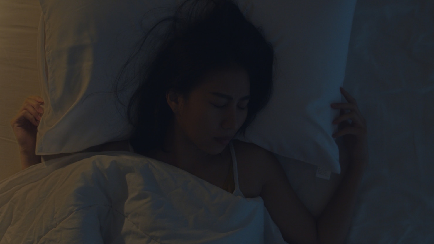 Person lying on a bed under a white comforter, resting on their back with arms spread out on either side of a white pillow. The room is dimly lit with soft, cool-toned lighting, creating a calm and peaceful nighttime atmosphere. The bedding is neatly arranged and the overall environment feels quiet and restful.