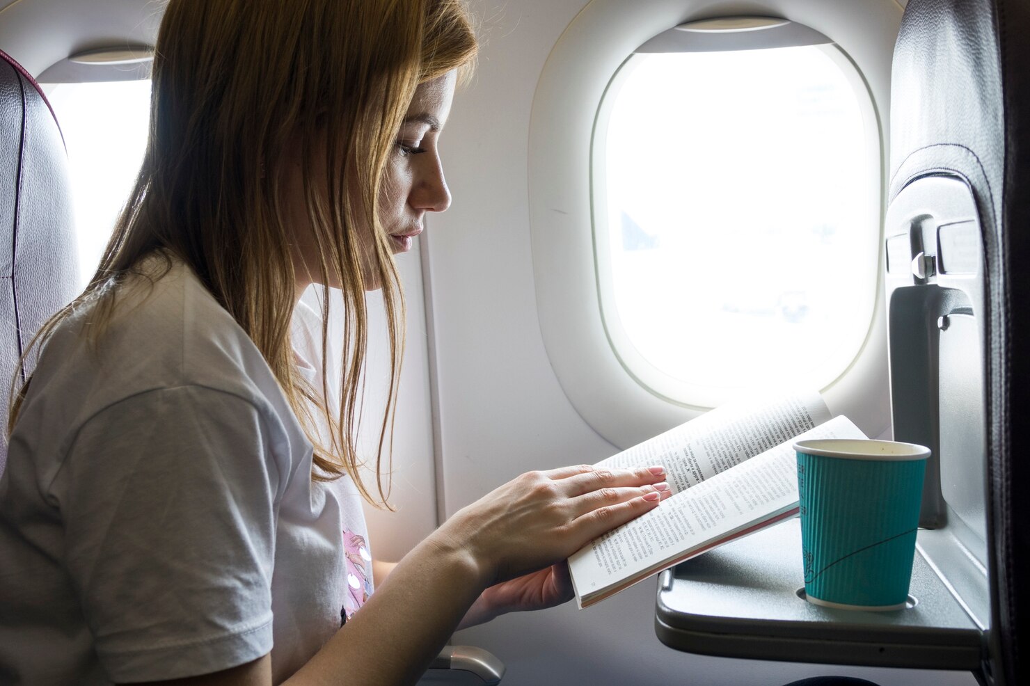Person with long hair sitting in an airplane seat by the window, reading a book held open on their lap. A turquoise paper cup sits on the tray table in front of them. The cabin is softly lit by daylight streaming through the window, creating a calm and focused atmosphere.