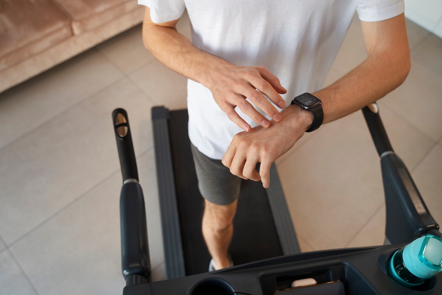 Person in a white t-shirt walking on a treadmill indoors, checking a smartwatch on their wrist. The environment is modern and clean with tiled floors and a sofa in the background. A water bottle is placed on the treadmill console, suggesting a focus on fitness and health tracking. The mood is active and attentive.