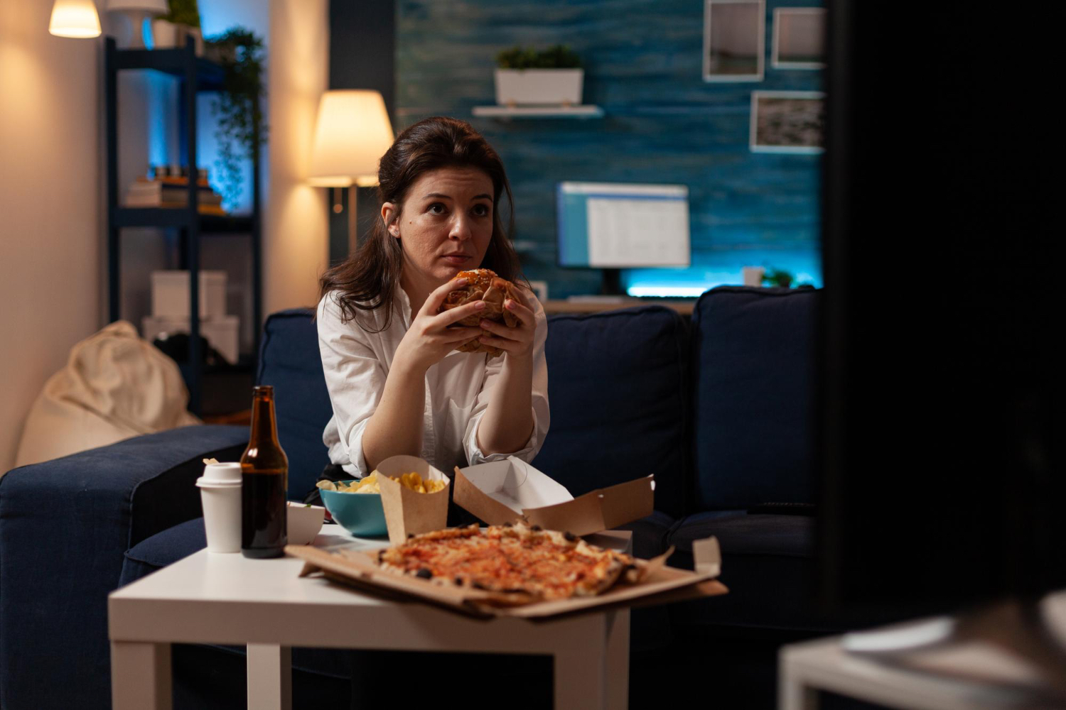 A woman sits on a sofa at night holding a burger while takeout food, pizza, chips, and drinks cover the coffee table in front of her.