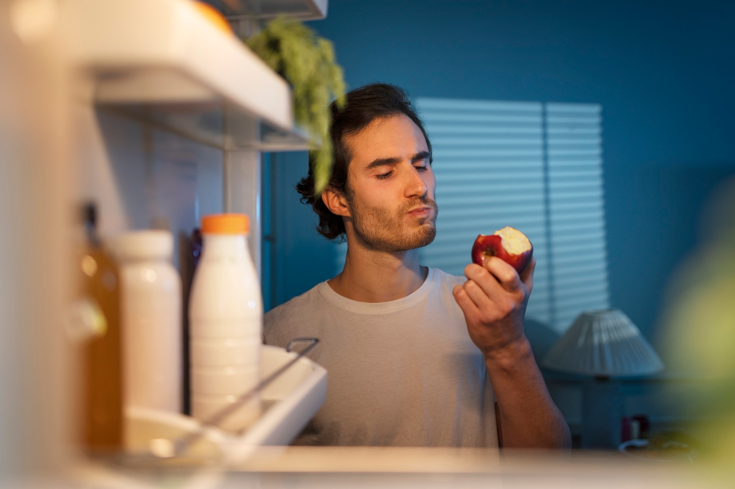 A man stands in front of an open refrigerator at night, taking a bite from a red apple while the fridge shelves glow beside him.