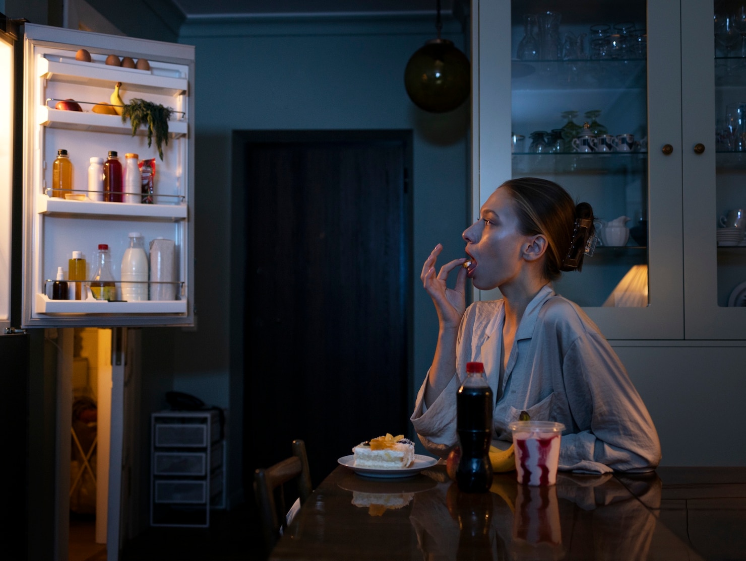 A woman sits at a kitchen table late at night eating dessert while an open refrigerator lights the dark room. A slice of cake, a soda bottle, and other snacks sit in front of her.