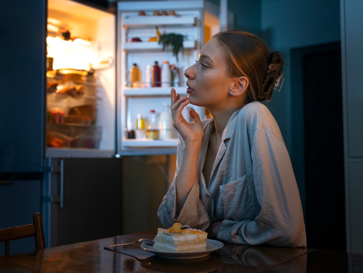 A woman in pajamas sits at a table late at night, looking toward an open refrigerator while a slice of cake rests on a plate in front of her.