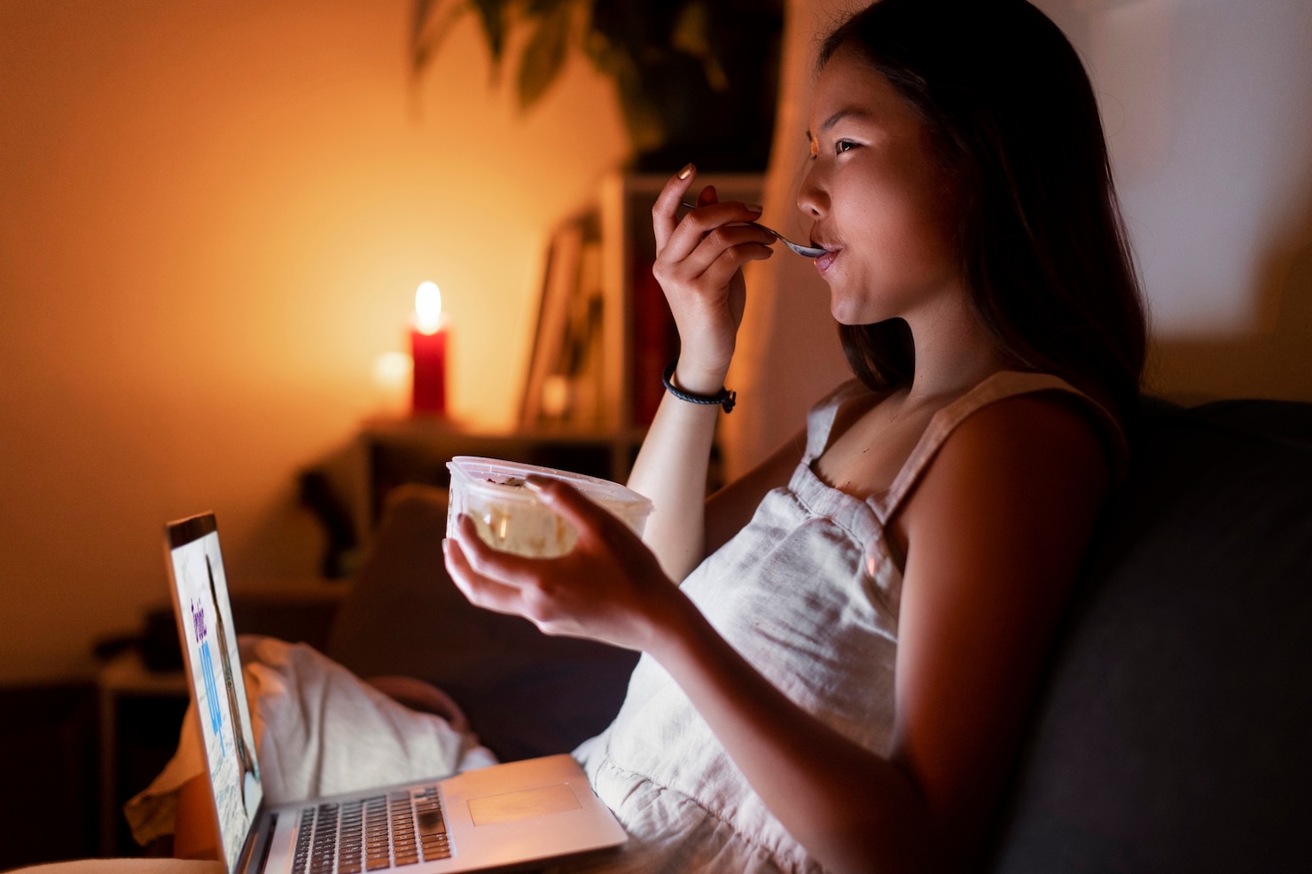 A woman sits on a couch at night eating from a plastic container with a spoon while a laptop glows on her lap and a candle burns in the background.