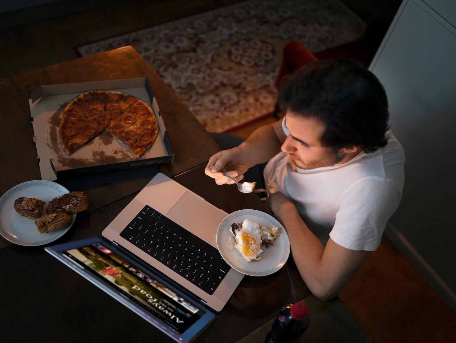 An overhead view shows a man eating dessert at a table with an open laptop, a partly eaten pizza in a box, and pastries on a plate beside him.