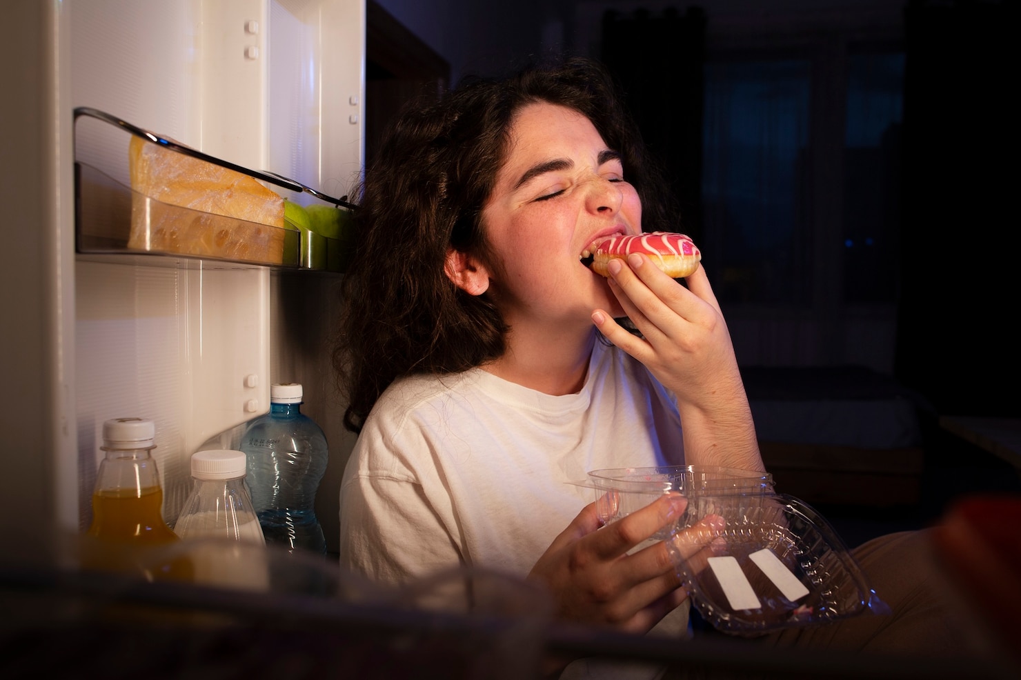 A woman sits beside an open refrigerator at night taking a bite from a pink-frosted donut while holding the empty pastry container.
