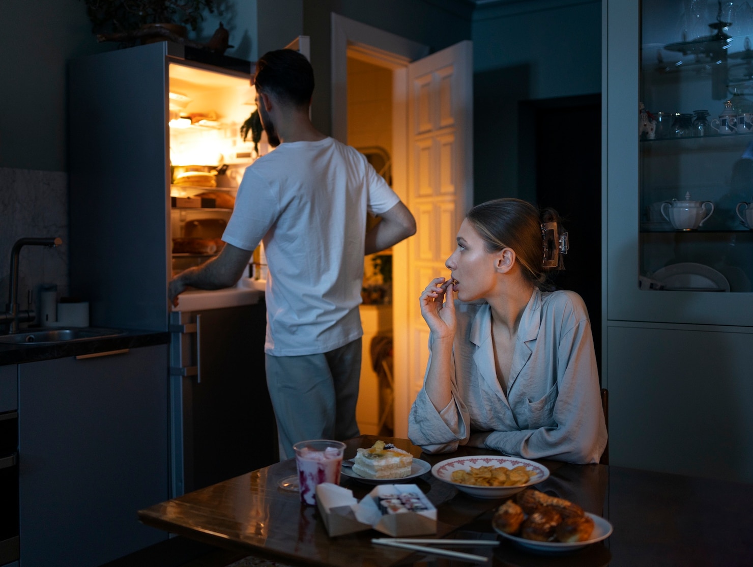 A woman sits at a kitchen table late at night with snacks and dessert while a man stands at the open refrigerator searching for more food.