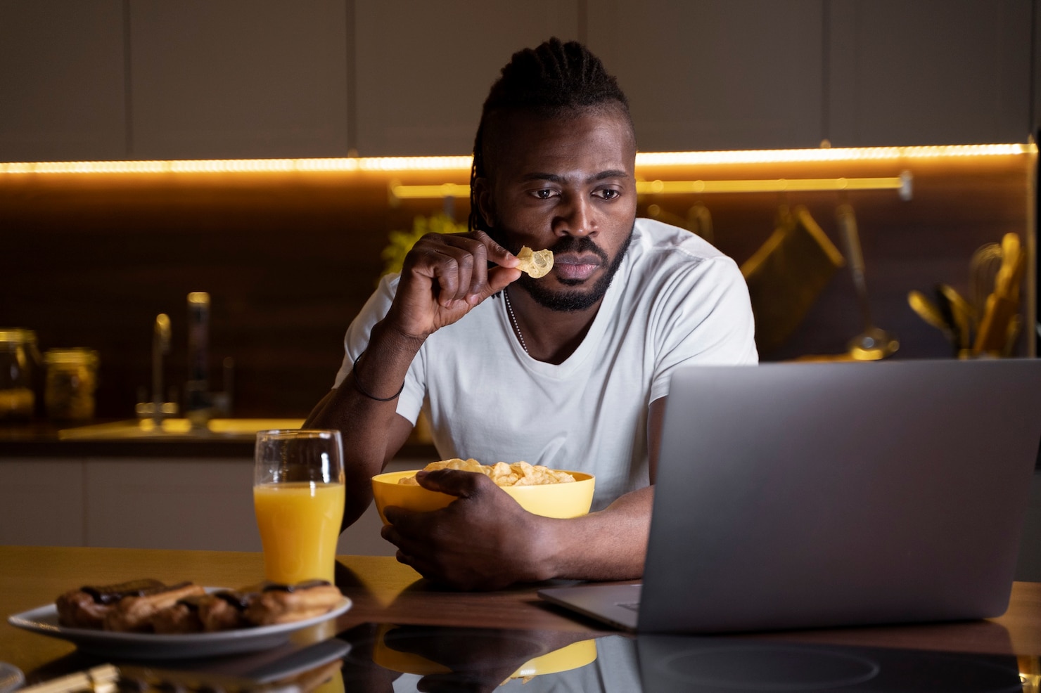 A man sits at a kitchen counter at night watching a laptop while holding a bowl of chips, with a glass of orange juice and pastries nearby.