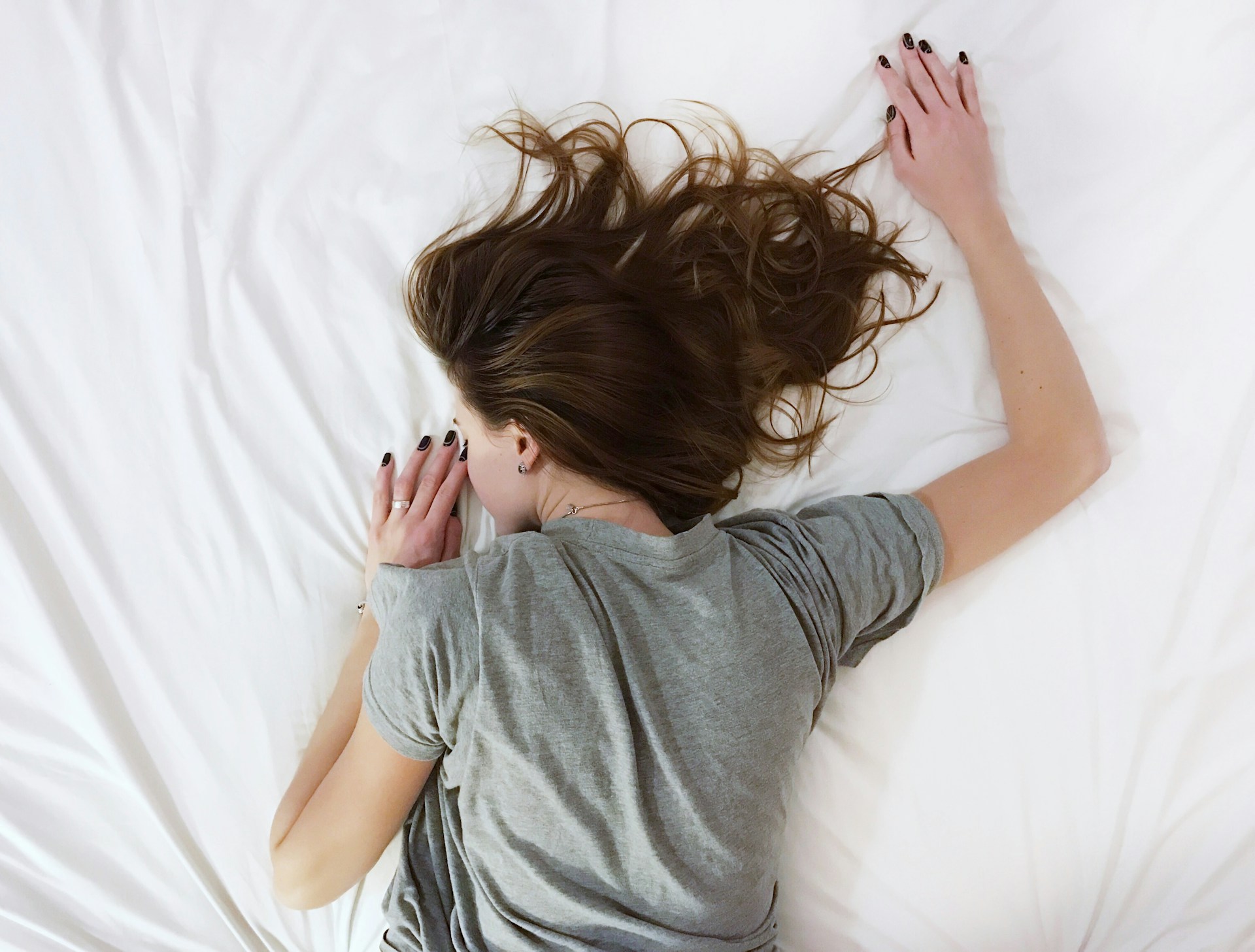 A woman with long brown hair lies face down on a bed with white sheets, wearing a gray t-shirt. Her arms are stretched out above her head and her hair is spread across the bed. The environment is calm and minimal, suggesting rest or exhaustion. The emotional tone feels peaceful and relaxed.