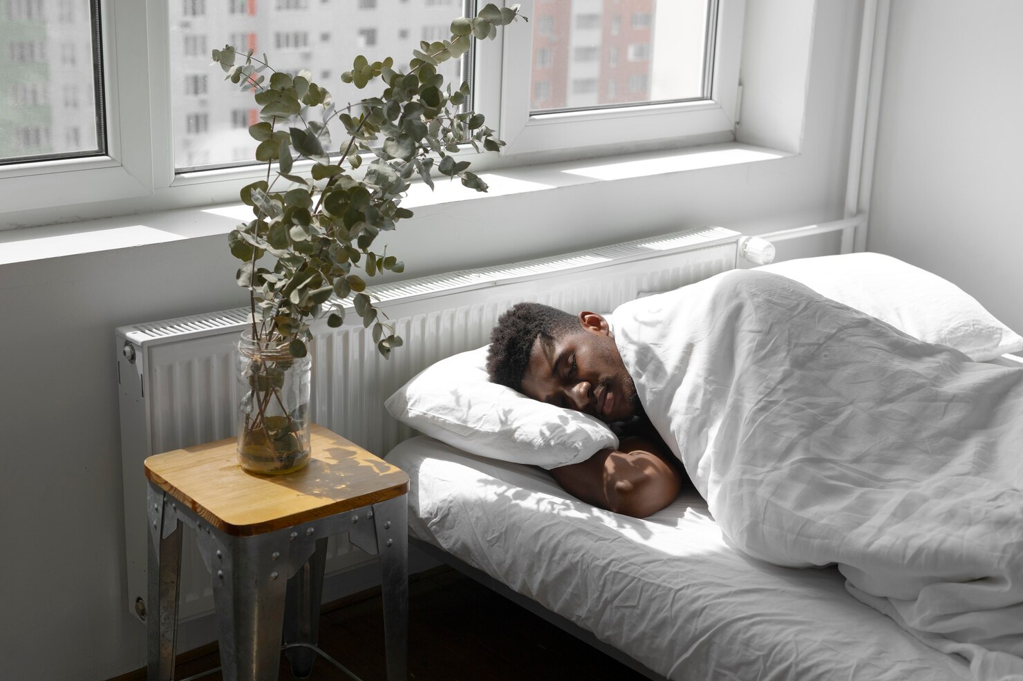 A man sleeps in bed beside a broad city window while strong daylight fills the room.
