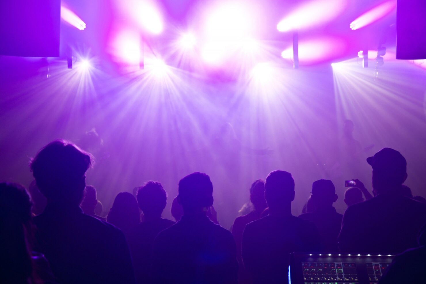 A crowd stands in a dark venue beneath intense purple stage lights, with beams of light spreading across the room.