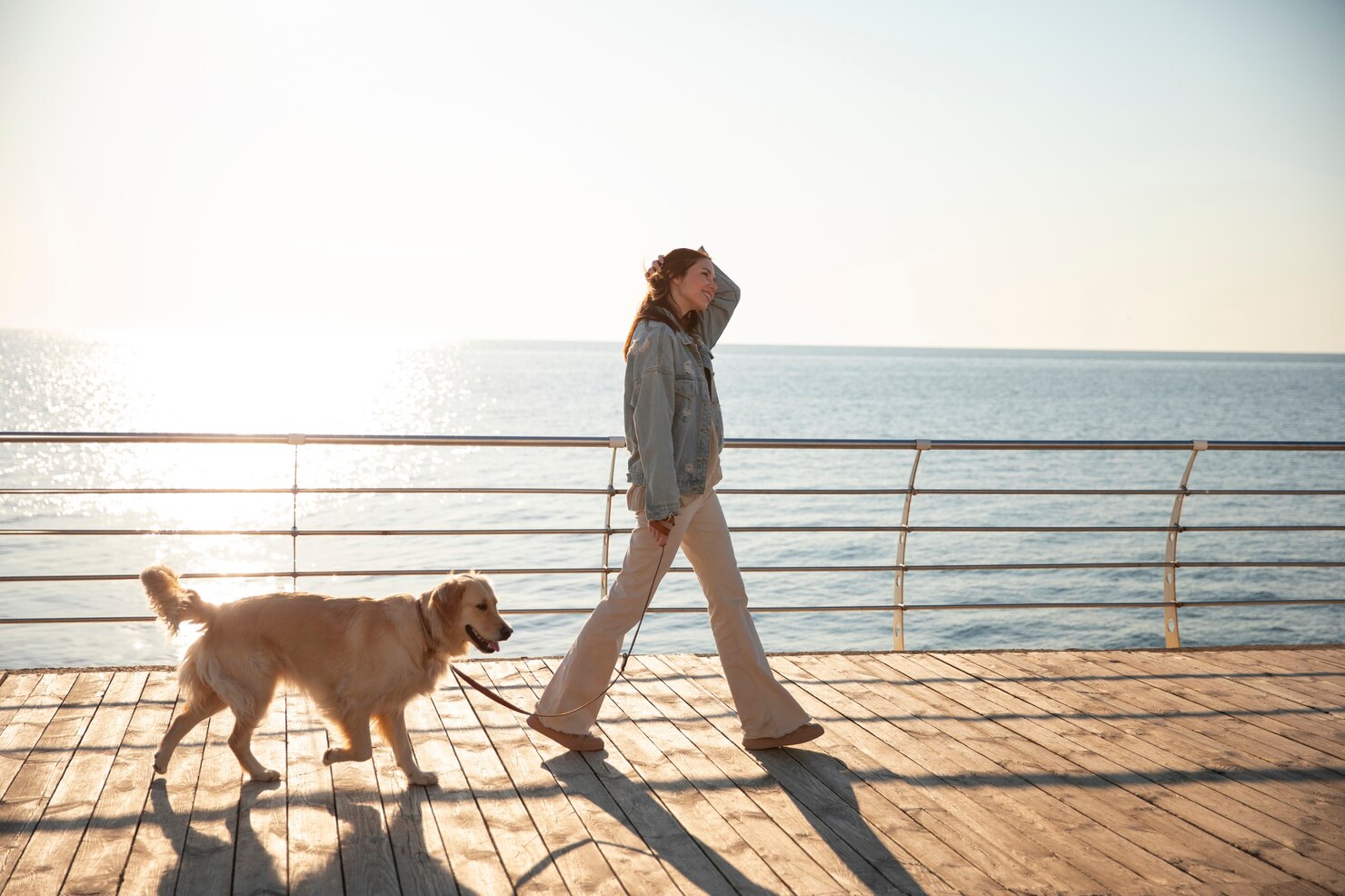 A woman walks a golden retriever along a sunlit boardwalk beside the water, backlit by bright morning light.