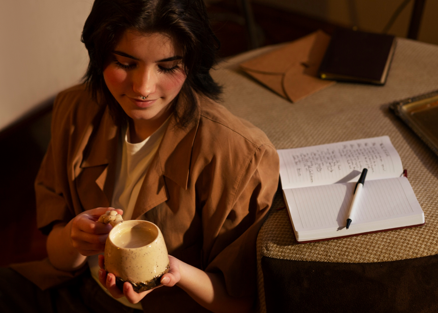 A person holds a large ceramic mug beside an open notebook and pen on a table in warm indoor light.