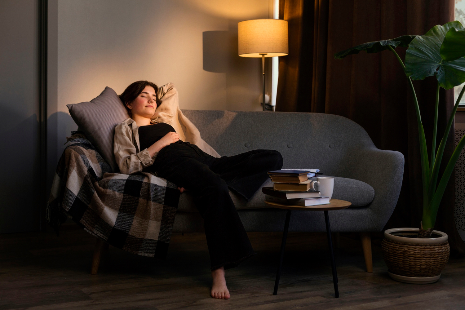 A woman naps on a gray sofa in a softly lit room, with a side table stacked with books and a white mug beside her.